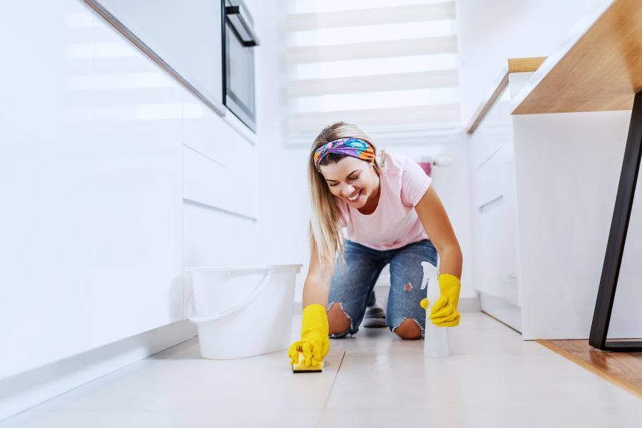 Dedicated smiling beautiful caucasian blond housewife with rubber gloves on kneeling and cleaning kitchen floor with detergent and sponge. Next to her is bucket with water.