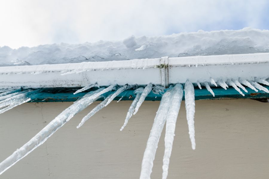 Icicles in all different shapes and sizes hanging down from the gutters as they overflow with a large chunk of ice flowing out of it