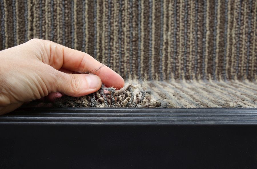 Person showing frayed carpet coming loose on stairstep, stair nosing or stair edging. High traffic staircase with commercial carpet damage by the edge, causing tripping hazard. Selective focus.