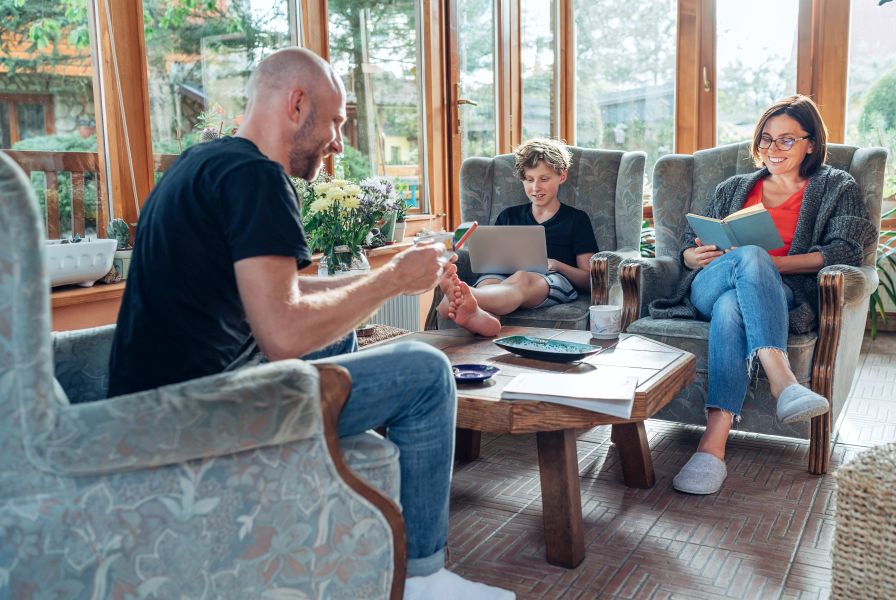 Mother, father and son family sitting together at Sunroom in cozy armchairs and reading books, using laptop or browsing a smartphone.